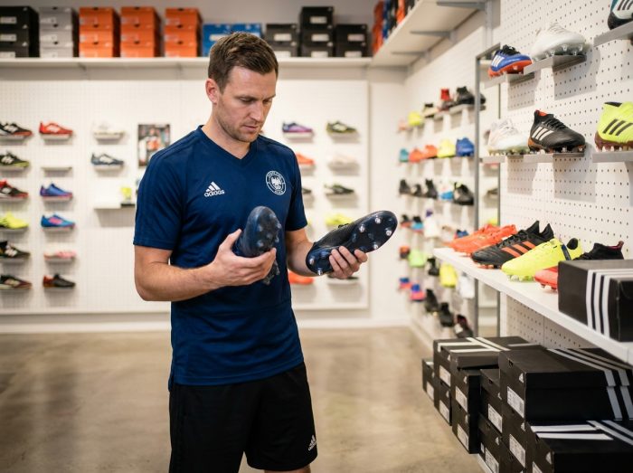 Footballeur comparant deux paires de crampons Adidas en magasin de sport pour choisir le modèle adapté à sa pratique hebdomadaire